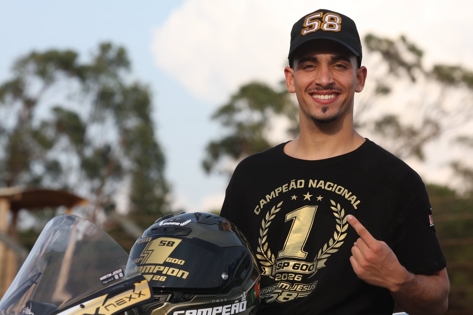 O Martim a sorrir, de boné com o 58, a apontar para o 1 da camisola de campeão, com o capacete ao lado